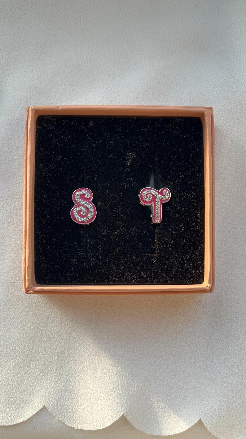 May include: A pink and silver zodiac earrings set in a box. The earrings are shaped like the zodiac signs for Sagittarius and Scorpio.