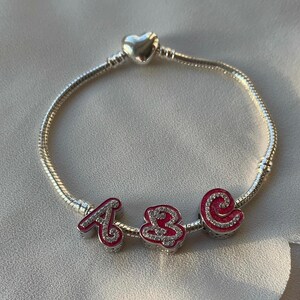 May include: A silver charm bracelet with a heart charm and three letter charms. The letter charms are pink with white glitter and spell out "ABC".