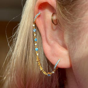 May include: A gold-colored earring with a curved design, featuring small blue and clear gemstones. The earring is worn on the ear, with a small gold hoop earring visible. Blonde hair is in the background.