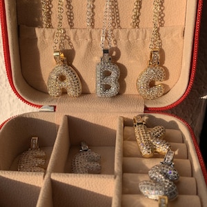 May include: A red jewellery box displaying several initial pendant necklaces. The pendants are encrusted with small, sparkling stones. The letters A, B, and C are visible, along with other letters. The necklaces have delicate chains.