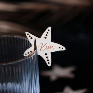 Può includere: Un segnaposto a forma di stella marina in legno con il nome "Kim" in corsivo. Il segnaposto, di colore chiaro, è attaccato al bordo di un bicchiere trasparente. Lo sfondo è scuro e sfocato.