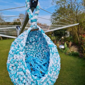 May include: A blue and white crocheted hanging basket filled with blue clothespins. The basket is hanging from a clothesline in a backyard.