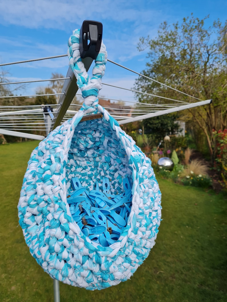 May include: A blue and white crocheted hanging basket filled with blue clothespins. The basket is hanging from a clothesline in a backyard.