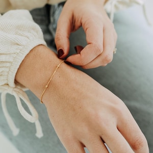 May include: A fine gold bracelet featuring small, evenly spaced beads. The bracelet is being adjusted on a wrist. The person is wearing a cream-coloured top and blue jeans.