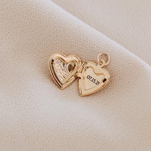 May include: A gold-toned heart-shaped locket pendant shown in an open position. The left interior side features an embossed floral pattern, while the right interior side displays the engraved date 07.15.21. A small circular jump ring is attached to the top.