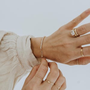 Peut inclure: Un bracelet doré orné d'une rangée de pierres claires et scintillantes. Le bracelet est relié par un fermoir en forme de trombone doré. Plusieurs bagues dorées ornent les doigts de la main portant le bracelet.