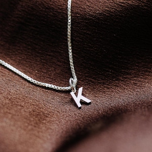 May include: A silver-toned necklace featuring a delicate box chain and a small, capital letter K pendant. The pendant is attached to the chain with a simple jump ring, showcasing a clean, metallic finish and a classic, minimalist letter design.