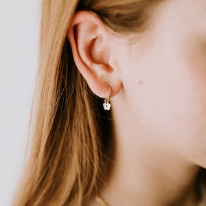 May include: A small gold hoop earring featuring a tiny, flat, five-petalled flower charm dangling from the bottom. The earring is worn in the earlobe, showcasing a polished metallic finish and a simple, delicate design with a circular hoop.