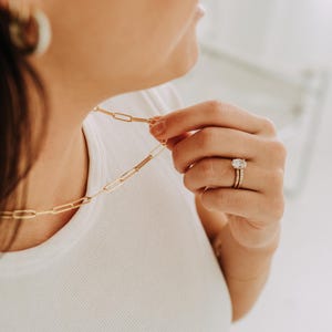 May include: A gold-colored paperclip chain necklace is held by a person wearing a white t-shirt. The person is also wearing a gold ring with a large, clear stone and a gold band. A gold hoop earring is visible.