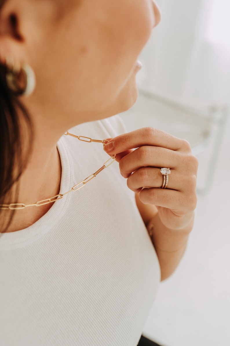 May include: A gold-colored paperclip chain necklace is held by a person wearing a white t-shirt. The person is also wearing a gold ring with a large, clear stone and a gold band. A gold hoop earring is visible.