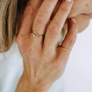 Peut inclure: Un ensemble de trois bagues fines en or portées aux doigts. Une bague présente un design en chevron ou en forme de V, une autre est un anneau simple avec une petite pierre blanche, et la troisième est un anneau en or lisse et uni.