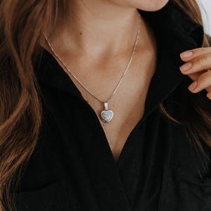 May include: A silver heart-shaped locket pendant suspended on a delicate silver chain. The locket features an engraved floral pattern on its front surface. The necklace is worn around the neck, resting against a black collared shirt.