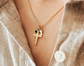 May include: A gold-colored necklace with a delicate chain and three charms. The charms include a cross, a small green gem, and a stylized letter. The necklace is worn against a beige shirt.