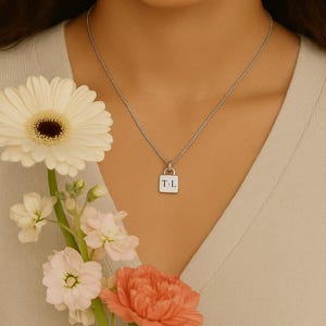 Peut inclure: Un collier en chaîne argent avec un pendentif carré sur lequel est inscrit "T.L.". Le collier est porté par une personne tenant un bouquet de fleurs blanches et roses.