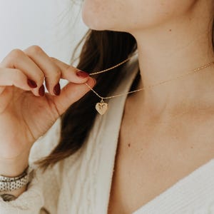 May include: A gold-toned heart-shaped locket pendant suspended from a delicate, thin gold-toned chain. The locket features an intricate, embossed floral or leaf-like pattern on its surface, reflecting light with a metallic finish.