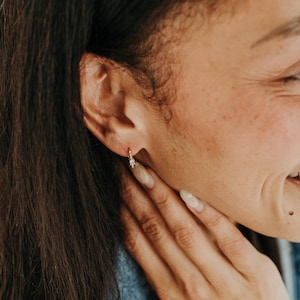 May include: A close-up of a person's ear wearing a small gold hoop earring with a small, decorative charm. The person is smiling, and their hand is gently touching their face. The earring is the focal point.