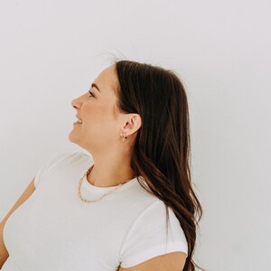 May include: A woman wearing a white t-shirt and gold jewelry, including a chain necklace and hoop earrings. Her dark brown hair is long and wavy. The background is a plain white wall.