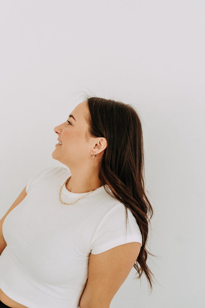 May include: A woman wearing a white t-shirt and gold jewelry, including a chain necklace and hoop earrings. Her dark brown hair is long and wavy. The background is a plain white wall.