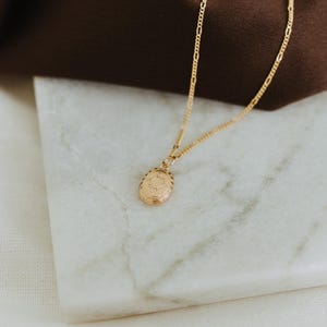 May include: A gold-toned necklace featuring a delicate chain and a small, oval-shaped pendant. The pendant has an embossed, intricate design on its front surface, suspended from a fine link chain that catches the light with its polished finish.