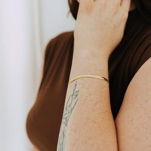 May include: A gold herringbone bracelet is worn on an arm with a floral tattoo. The person is wearing a brown top and several gold rings. The bracelet is a delicate piece of jewellery.