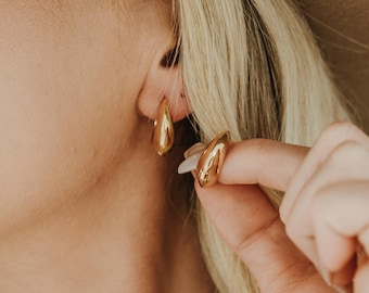 May include: Gold teardrop-shaped earrings. The earrings have a smooth, polished surface and a curved design. The image shows the earrings being worn, with one earring in focus and the other slightly blurred in the background. The earrings are a shiny gold color.