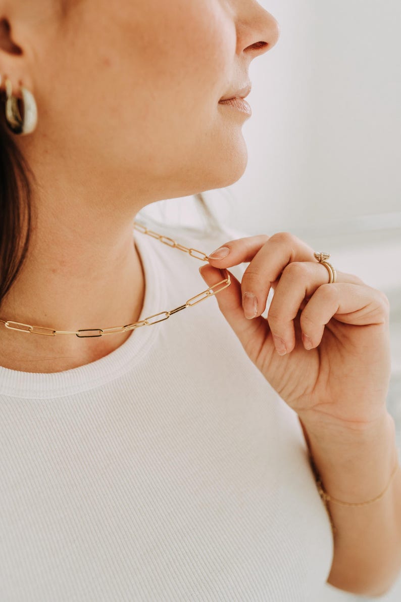 May include: A gold-colored paperclip chain necklace is held by a person wearing a white ribbed t-shirt. The necklace is made of elongated rectangular links. A gold ring and bracelet are also visible.