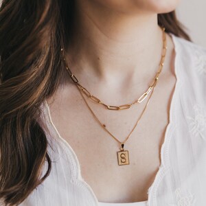 May include: A gold-colored necklace set featuring two layered chains. The top chain has large rectangular links, while the lower chain holds a square pendant with the letter "S". The jewelry is worn over a white top.