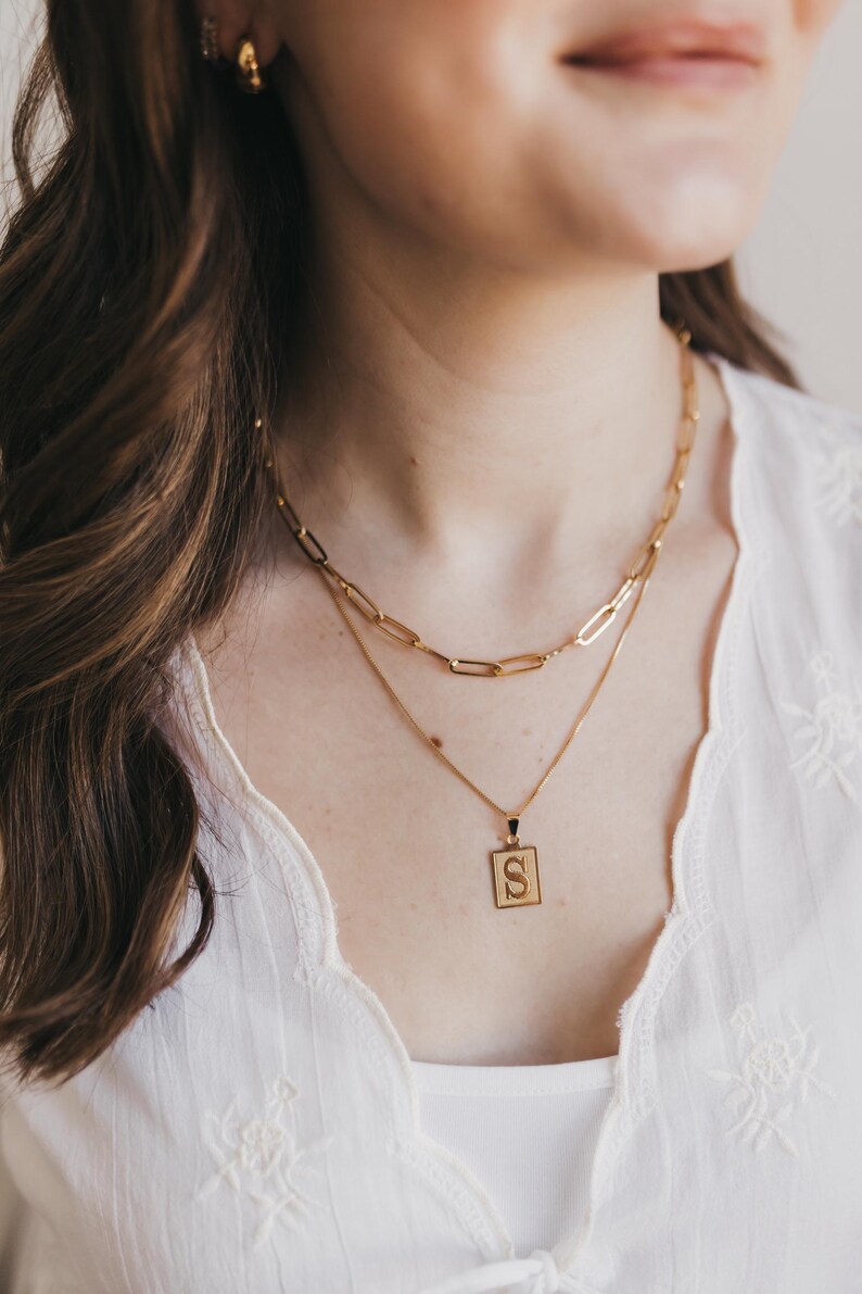 May include: A gold-colored necklace set featuring two layered chains. The top chain has large rectangular links, while the lower chain holds a square pendant with the letter "S". The jewelry is worn over a white top.