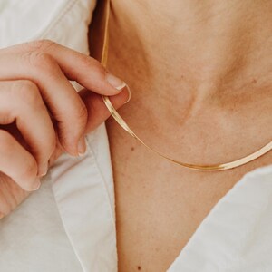 May include: A gold-toned herringbone chain necklace with a flat, flexible link design. The necklace rests against the collarbone, featuring a polished, reflective surface that catches the light. The chain is thin and smooth, creating a continuous line.