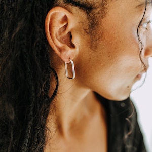 Peut inclure: Boucles d'oreilles créoles rectangulaires argentées. Les boucles d'oreilles sont portées à l'oreille d'une personne, l'accent étant mis sur les bijoux. Les boucles d'oreilles ont un design simple et moderne. La personne porte un haut noir.
