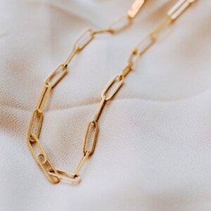 May include: A gold-colored chain necklace with a paperclip link design. The necklace is displayed on a white, textured surface. The links are elongated and rectangular, creating a modern and minimalist aesthetic. The chain is a bright, shiny gold.