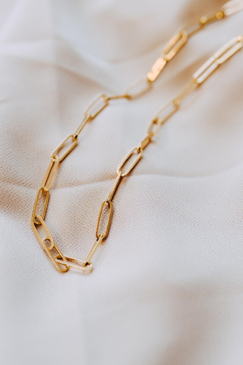May include: A gold-colored chain necklace with a paperclip link design. The necklace is displayed on a white, textured surface. The links are elongated and rectangular, creating a modern and minimalist aesthetic. The chain is a bright, shiny gold.