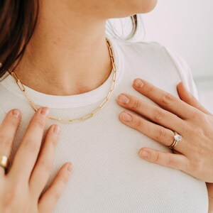 May include: A gold-colored chain necklace with a rectangular link design. The necklace is worn on a person wearing a white ribbed t-shirt. A gold ring and earrings are also visible.
