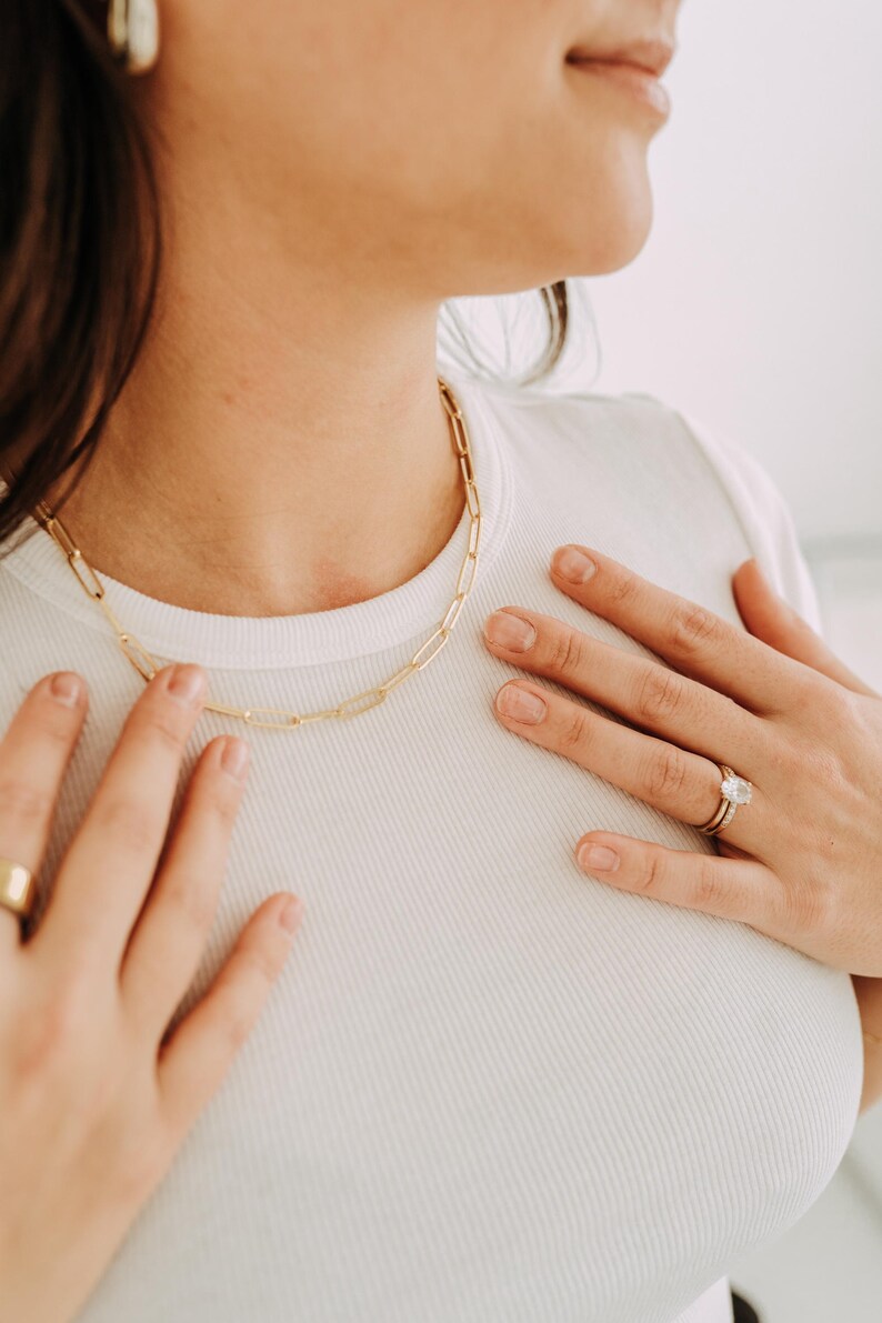 May include: A gold-colored chain necklace with a rectangular link design. The necklace is worn on a person wearing a white ribbed t-shirt. A gold ring and earrings are also visible.