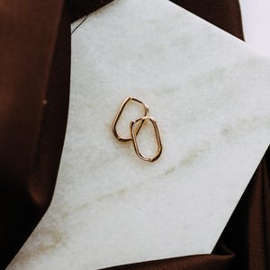 May include: A pair of small, gold-toned hoop earrings with an elongated, rectangular shape and rounded corners. The earrings feature a polished metallic finish and a simple hinged closure mechanism, presented as a matching set of two individual pieces.