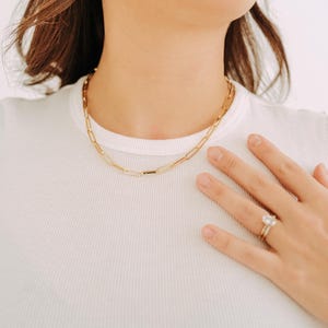 May include: A gold-colored chain necklace with a paperclip link design. The necklace is worn on a person wearing a white t-shirt. A gold ring with a large, clear stone is on the person's finger.