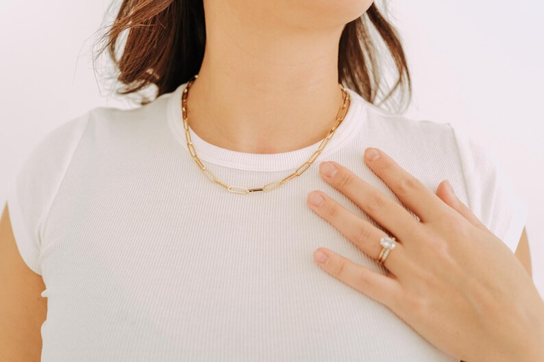 May include: A gold-colored chain necklace with a paperclip link design. The necklace is worn on a person wearing a white t-shirt. A gold ring with a large, clear stone is on the person's finger.