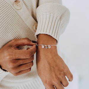 Peut inclure: Un bracelet à chaîne en argent avec trois petits pendentifs : deux cœurs gravés des lettres E et H, et un pendentif circulaire central orné d'une pierre bleu clair. Le bracelet est porté au poignet.