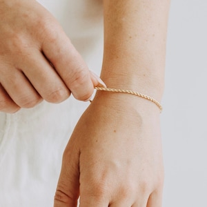 May include: A gold rope chain bracelet is being fastened on a wrist. The bracelet has a delicate, twisted rope design. The skin tone is fair, and the background is a soft, neutral colour. The bracelet's length is approximately 17 cm.