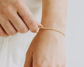 May include: A gold-colored rope chain bracelet is being clasped on a wrist. The bracelet is thin and delicate, with a twisted rope design. The skin tone is fair, and the background is a soft, neutral color.