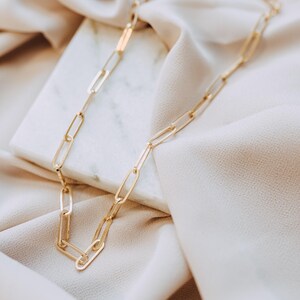 May include: A gold-colored chain necklace with elongated rectangular links. The necklace is displayed on a white marble surface and beige fabric, creating a soft, elegant aesthetic. The chain's design is simple yet stylish.