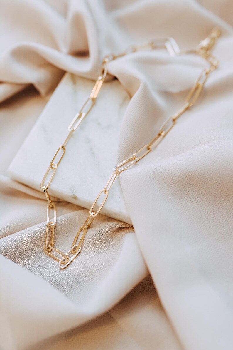 May include: A gold-colored chain necklace with elongated rectangular links. The necklace is displayed on a white marble surface and beige fabric, creating a soft, elegant aesthetic. The chain's design is simple yet stylish.