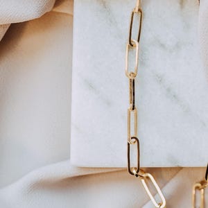 May include: A gold-colored chain necklace with elongated rectangular links. The necklace is displayed on a white marble surface, with a soft, off-white fabric in the background. The chain is a simple, elegant design.