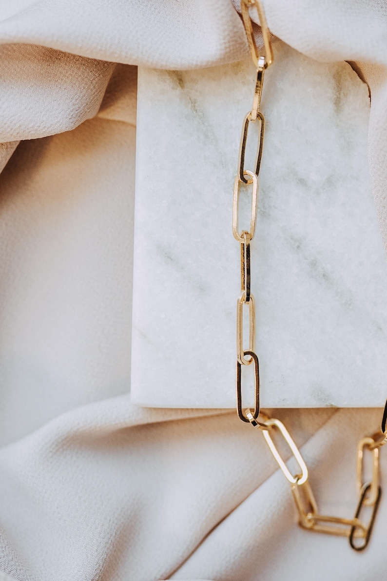 May include: A gold-colored chain necklace with elongated rectangular links. The necklace is displayed on a white marble surface, with a soft, off-white fabric in the background. The chain is a simple, elegant design.