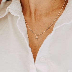 Peut inclure: Un collier à chaîne fine de couleur dorée avec un petit pendentif simple en forme de lettre majuscule K. Le collier est porté autour du cou, reposant sur une chemise blanche à col avec des boutons visibles et une finition en tissu texturé.