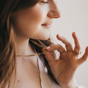 May include: A gold-colored paperclip chain necklace is held up by a person's fingers. Several gold rings adorn the fingers. A gold bracelet is on the wrist. The jewelry is displayed against a soft, neutral background.