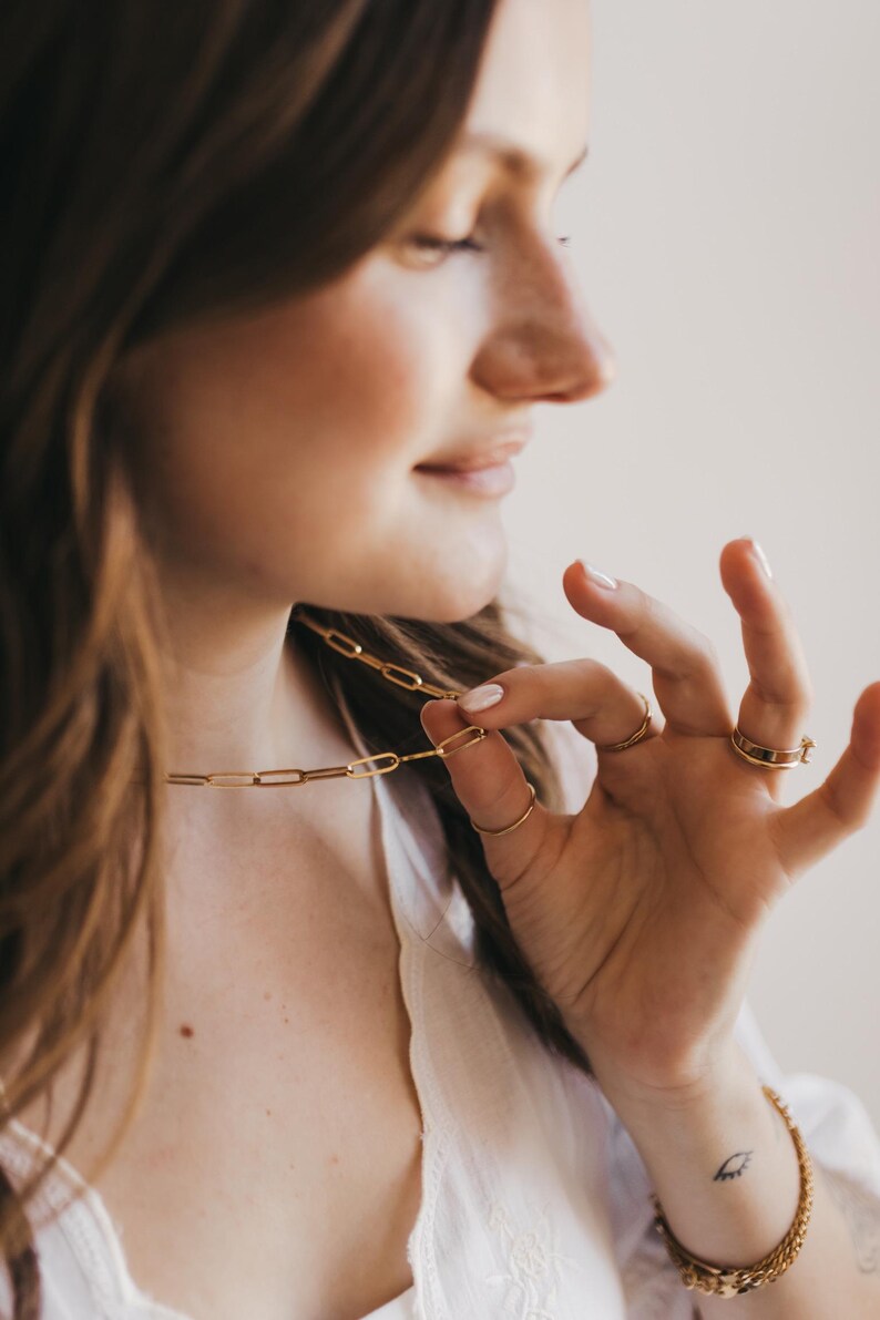 May include: A gold-colored paperclip chain necklace is held up by a person's fingers. Several gold rings adorn the fingers. A gold bracelet is on the wrist. The jewelry is displayed against a soft, neutral background.