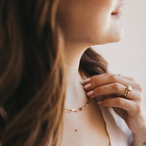 May include: A gold chain necklace with a rectangular link design. The necklace is worn around the neck. A gold ring with a large, square-cut stone is on the finger.