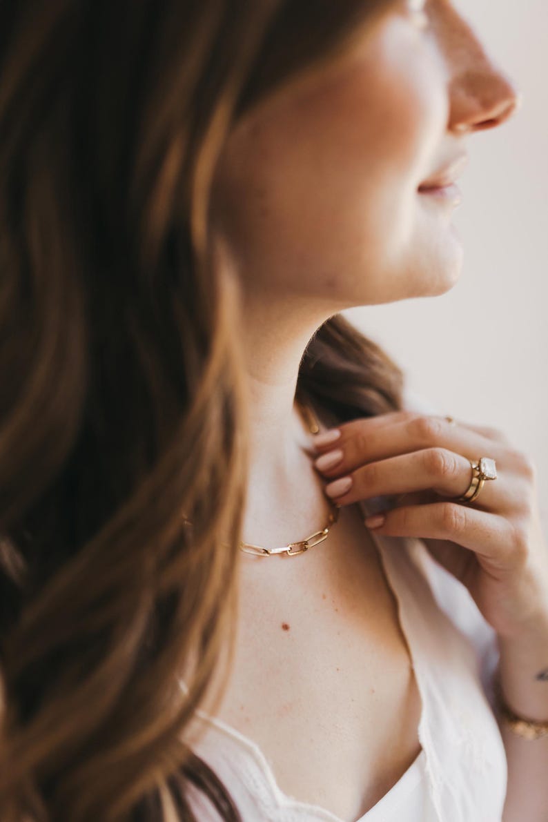 May include: A gold chain necklace with a rectangular link design. The necklace is worn around the neck. A gold ring with a large, square-cut stone is on the finger.