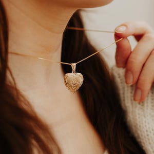 May include: A gold-toned heart-shaped locket pendant suspended on a delicate matching gold-toned chain. The locket features an intricate embossed floral or scrollwork border surrounding a central shield-like design with a small heart detail.